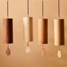 Four wooden cylinder wind chimes with teardrop-shaped metal striker weights hanging from black cords against a warm beige background.