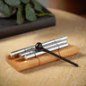 A set of silver metal cylindrical accessories with a black spherical handle and black cord, displayed on a small wooden stand on a beige tabletop.
