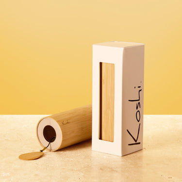 A bamboo wind instrument case standing upright next to its open cylindrical bamboo case, with a small wooden tag hanging from the open end. The white box has the brand name 'Koshi' printed vertically in black.