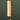 A natural wood wind chime hanging from a thin tree branch against a dark green background. The chime features a cylindrical wooden body with a small teardrop-shaped wooden striker at the bottom, suspended by a black cord with brass-colored hardware.