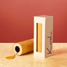 A bamboo cylindrical product with a black circular end and a small gold tag, shown next to its off-white rectangular packaging box with a vertical window and black handwritten text, set against a warm terracotta background and beige surface.