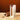 A white rectangular box with a clear window displaying a wooden Koshi rice stick, standing next to a tipped-over wooden container on a beige stone surface against a warm brown background. The brand name 'Koshi' is printed on both the box and the lid.