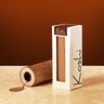A white rectangular box with a clear window displaying a wooden Koshi rice stick, standing next to a tipped-over wooden container on a beige stone surface against a warm brown background. The brand name 'Koshi' is printed on both the box and the lid.