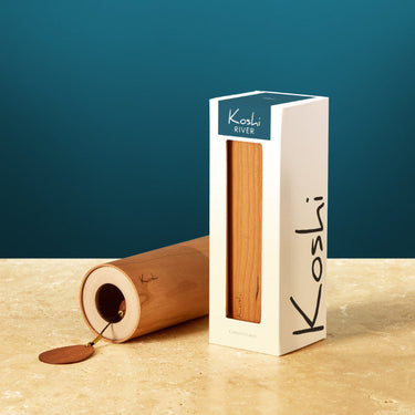 A Koshi rice wine stopper and its packaging on a beige stone surface against a dark teal background. The stopper, made of natural Koshi rice, is shown lying on its side with a small piece of rice visible inside, next to a white box with a clear window displaying the wood grain.