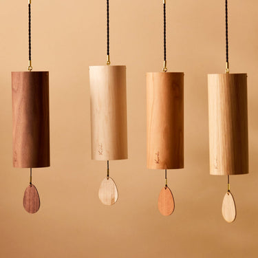 Four wooden cylinder wind chimes with teardrop-shaped metal striker weights hanging from black cords against a warm beige background.