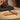 A silver metal flute-like instrument with a black mallet rests on a small bamboo holder on a wooden surface, with a dark rectangular object and a blurred wooden decorative object in the background.