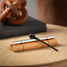 A silver metal flute-like instrument with a black mallet rests on a small bamboo holder on a wooden surface, with a dark rectangular object and a blurred wooden decorative object in the background.