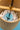Close-up of a wooden hanging ornament or wind spinner with a blue glass disc, metal rods, and a black string inside a cylindrical wooden frame.