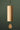 A natural wood wind chime hanging from a thin tree branch against a dark green background. The chime features a cylindrical wooden body with a small teardrop-shaped wooden striker at the bottom, suspended by a black cord with brass-colored hardware.