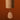A wooden wind chime with a cylindrical body and a teardrop-shaped hanging striker, both in a warm natural wood finish, set against a solid dark orange-brown background.