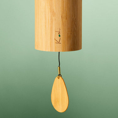 A bamboo wind chime with a hanging wooden striker and black cord, shown against a soft green gradient background.