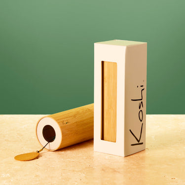 A rectangular cream-colored box with a vertical window displaying a light wood insert, standing next to a matching cylindrical wooden box lying on its side. The cream box has the brand name 'Kosli' written vertically in black.