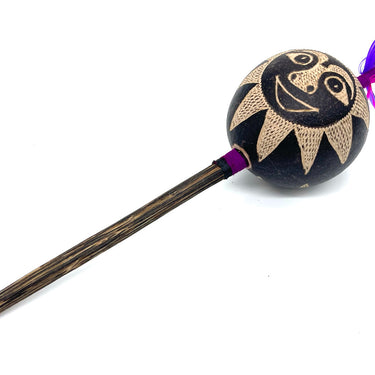 A handcrafted wooden mace or club with a round black head decorated with a beige sun face design and purple and pink feathers, set against a plain white background.