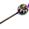 A handcrafted wooden mace or club with a round black head decorated with a beige sun face design and purple and pink feathers, set against a plain white background.
