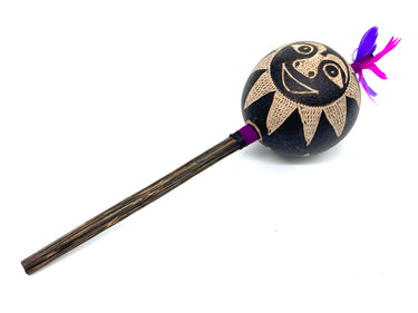 A handcrafted wooden mace or club with a round black head decorated with a beige sun face design and purple and pink feathers, set against a plain white background.
